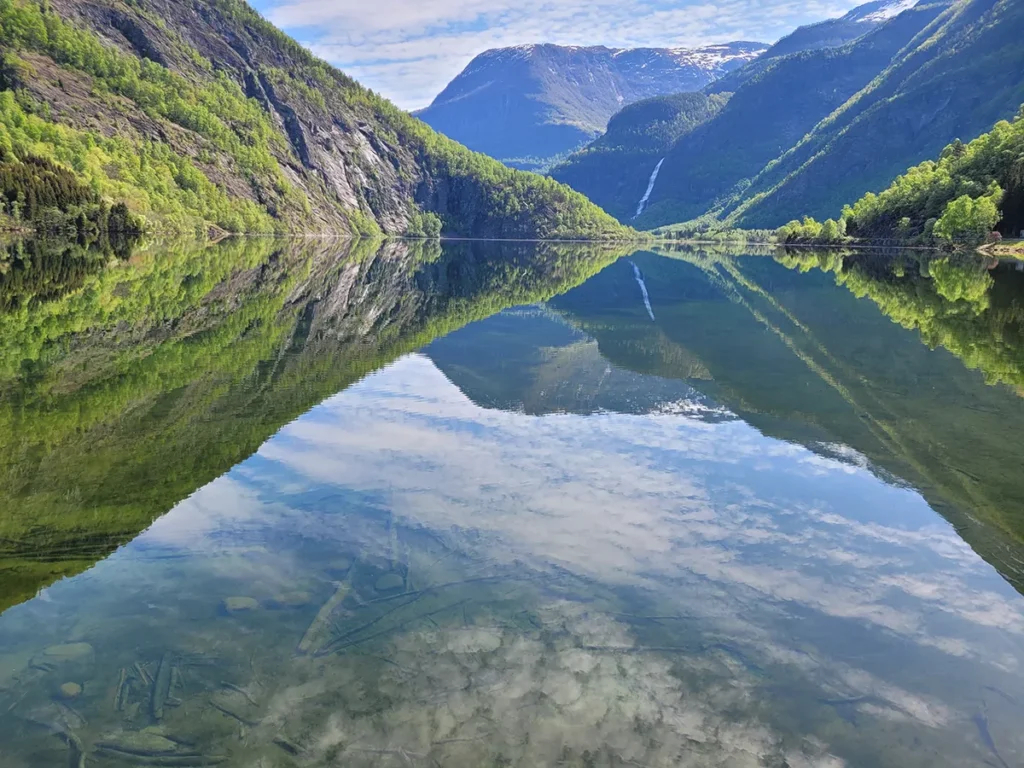 Side angle of Eidsvatnet lake showcasing the rocky shoreline, lush grass, and mountain ranges under a cloudy sky.
