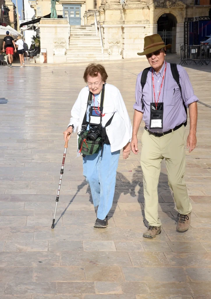 A married tourist couple walking down a historic narrow street with traditional colorful balconies in Valletta, Malta.